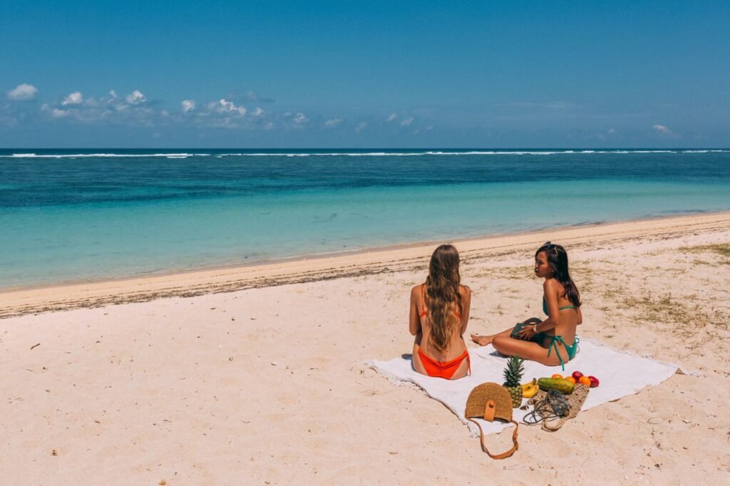 Two women in swimsuits enjoying a sunny day on a tropical beach with clear blue waters.